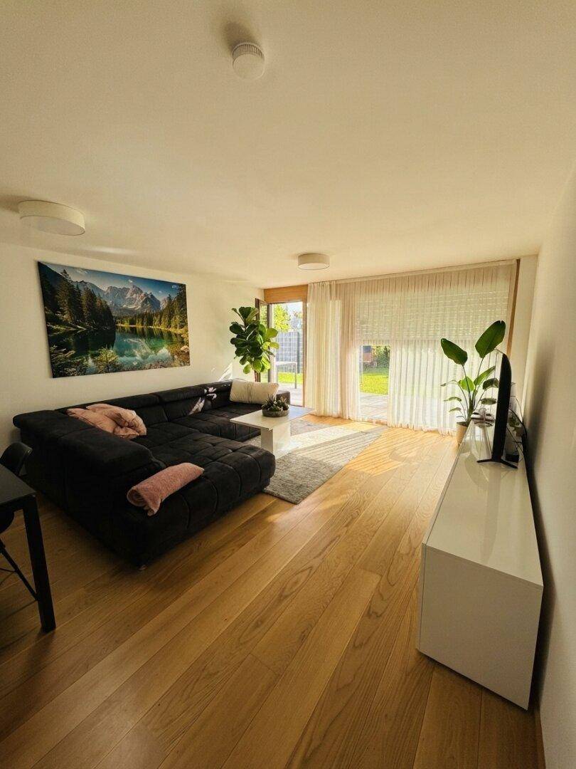 Geräumiges Wohnzimmer mit dunklem Ecksofa, Fernseher und großen Fenstern.