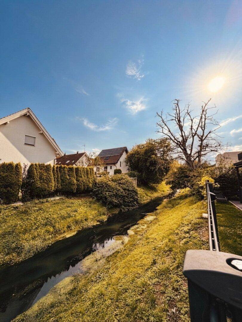 Malerischer Bachlauf neben Wohnhäusern mit üppigem Grün unter strahlend blauem Himmel.