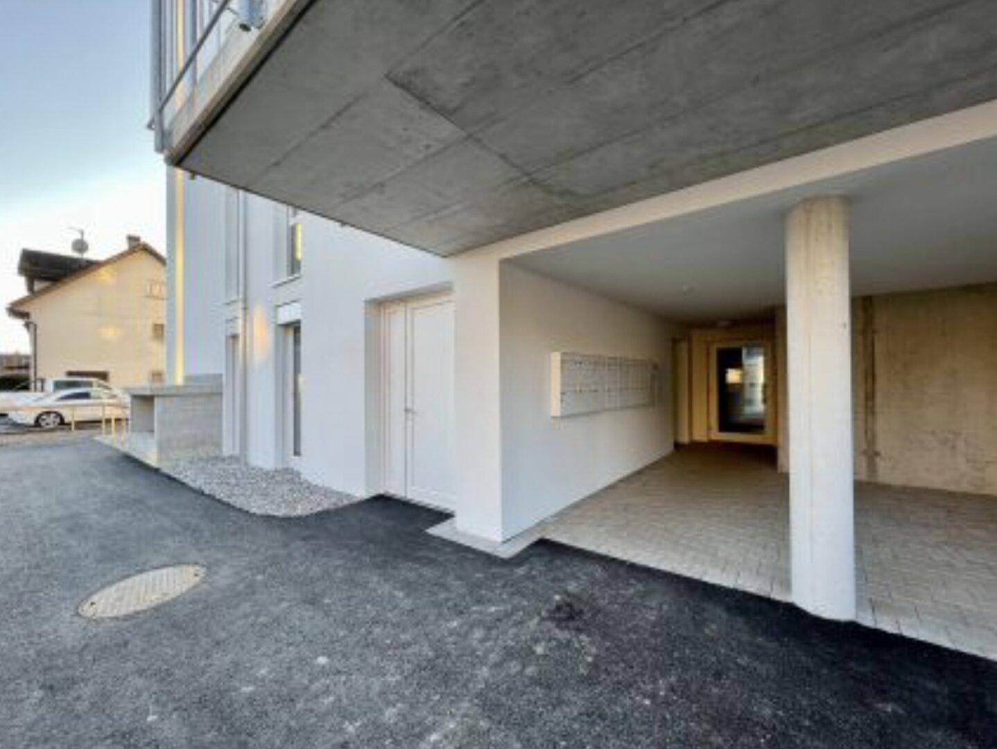 Eingangsbereich eines modernen Gebäudes mit überdachtem Zugang und Briefkästen, daneben ein Parkplatz.