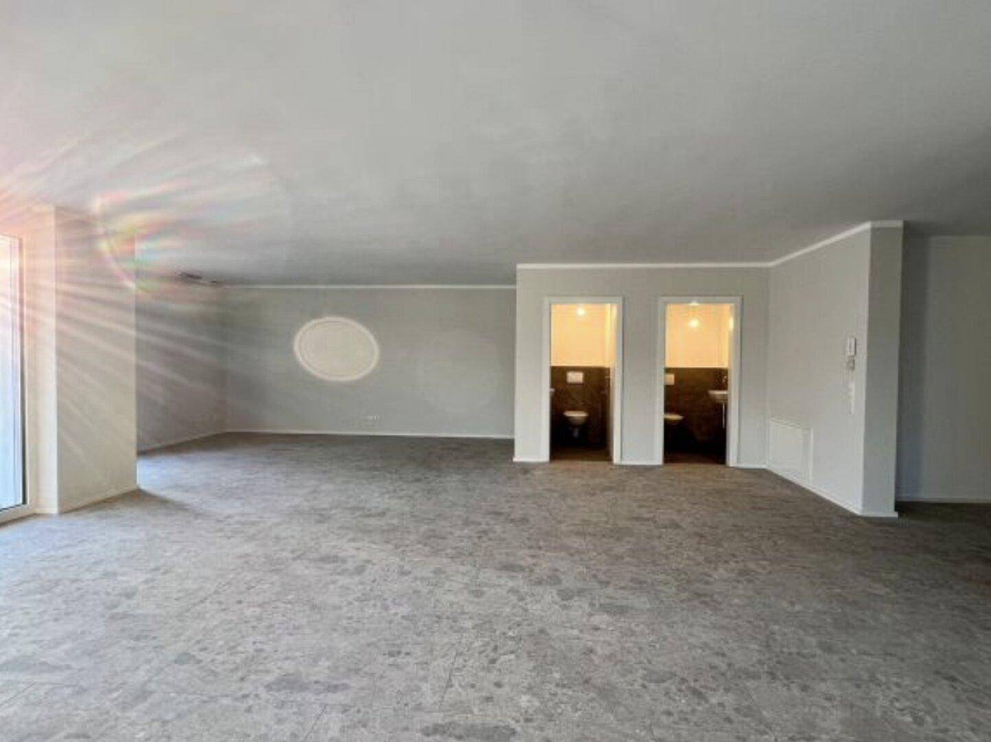 Großer, leerer Raum mit Fliesenboden und zwei sichtbaren Toilettenräumen im Hintergrund.