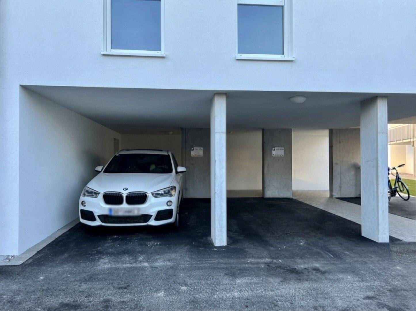 Überdachter Stellplatz unter dem Gebäude mit einem geparkten weißen Auto und moderner Architektur.