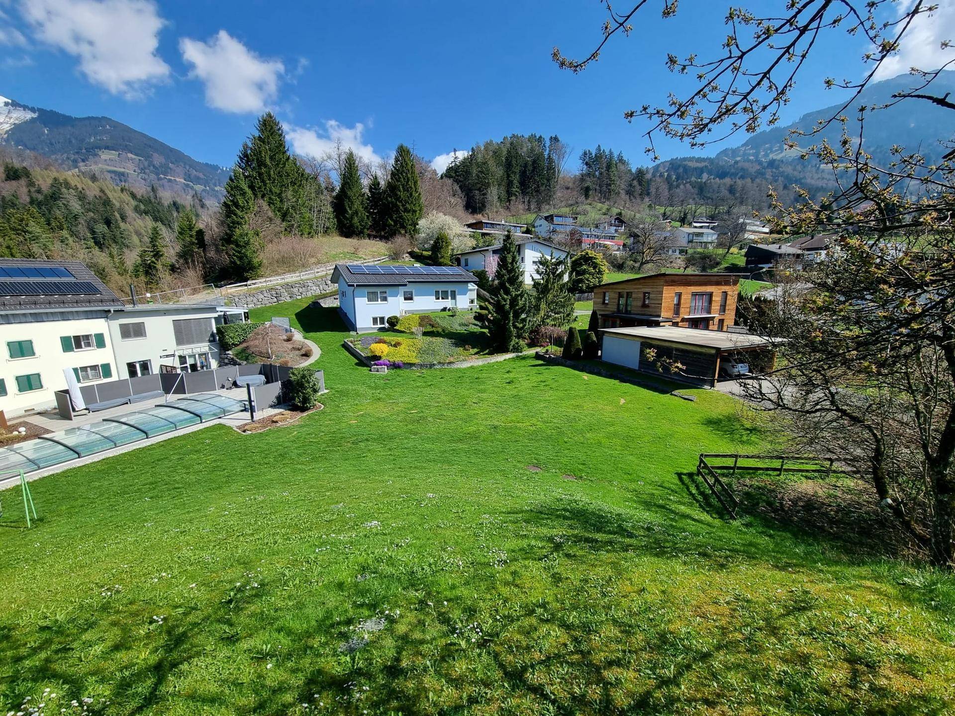 Weitläufiger Blick über eine Wohnsiedlung mit grünen Gärten und bewaldeten Bergen im Hintergrund.