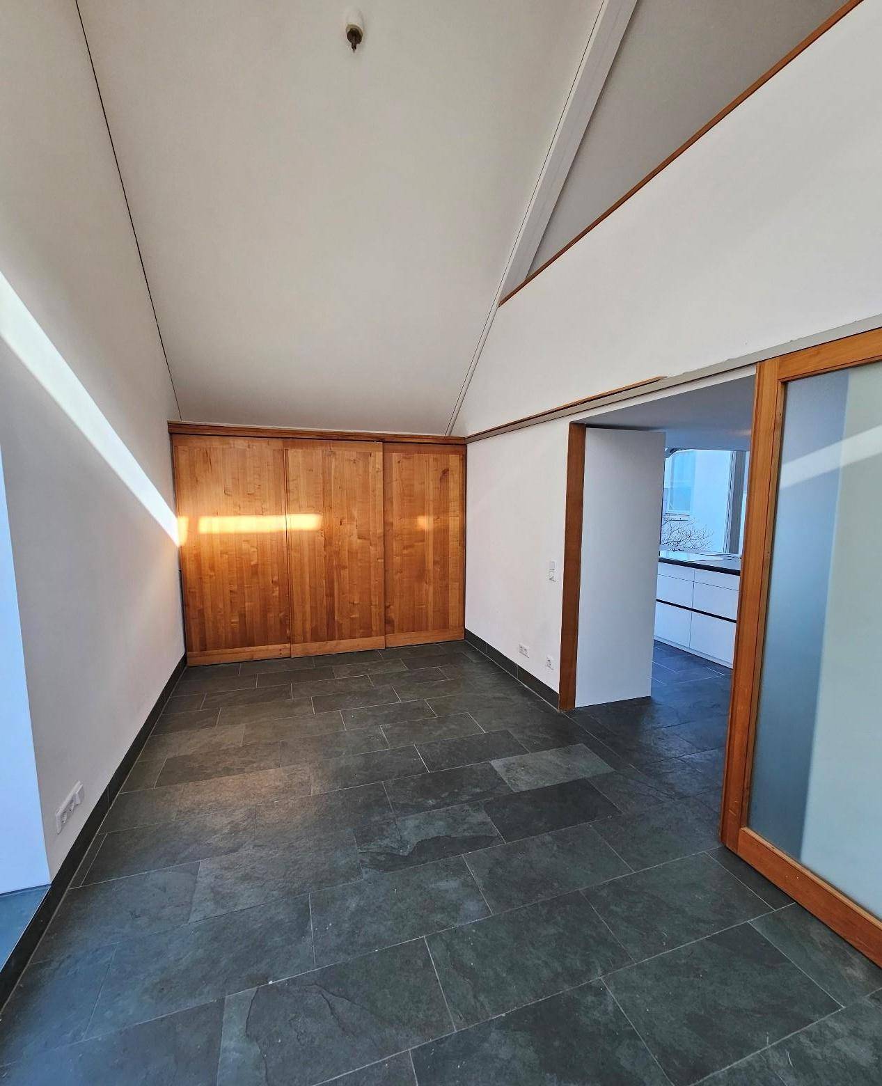 Heller Raum mit dunklem Fliesenboden und Holzwand, offen zu einem weiteren Bereich.