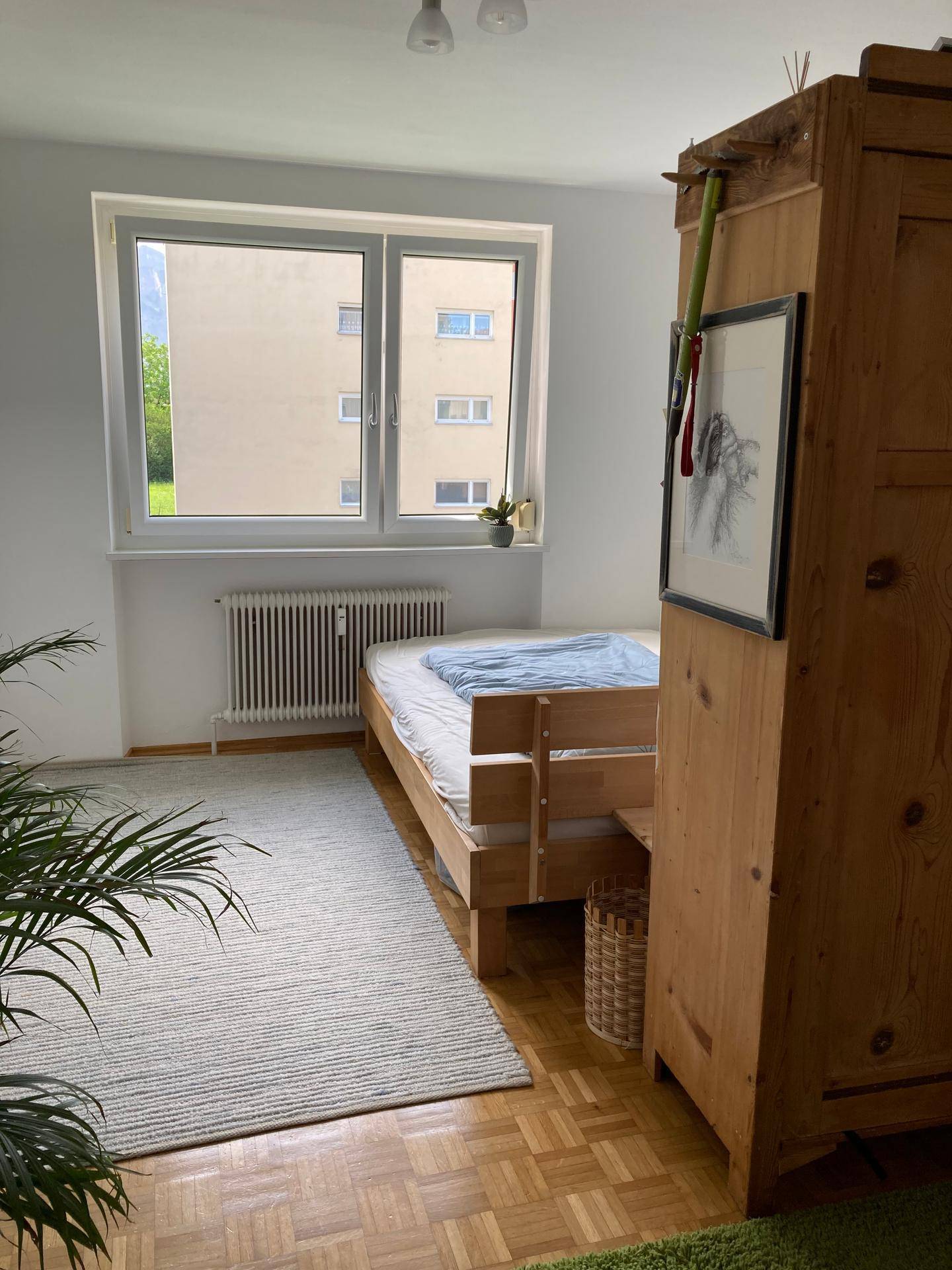 Helles Schlafzimmer mit Einzelbett, Parkettboden und Fenster mit Blick auf Nachbargebäude.