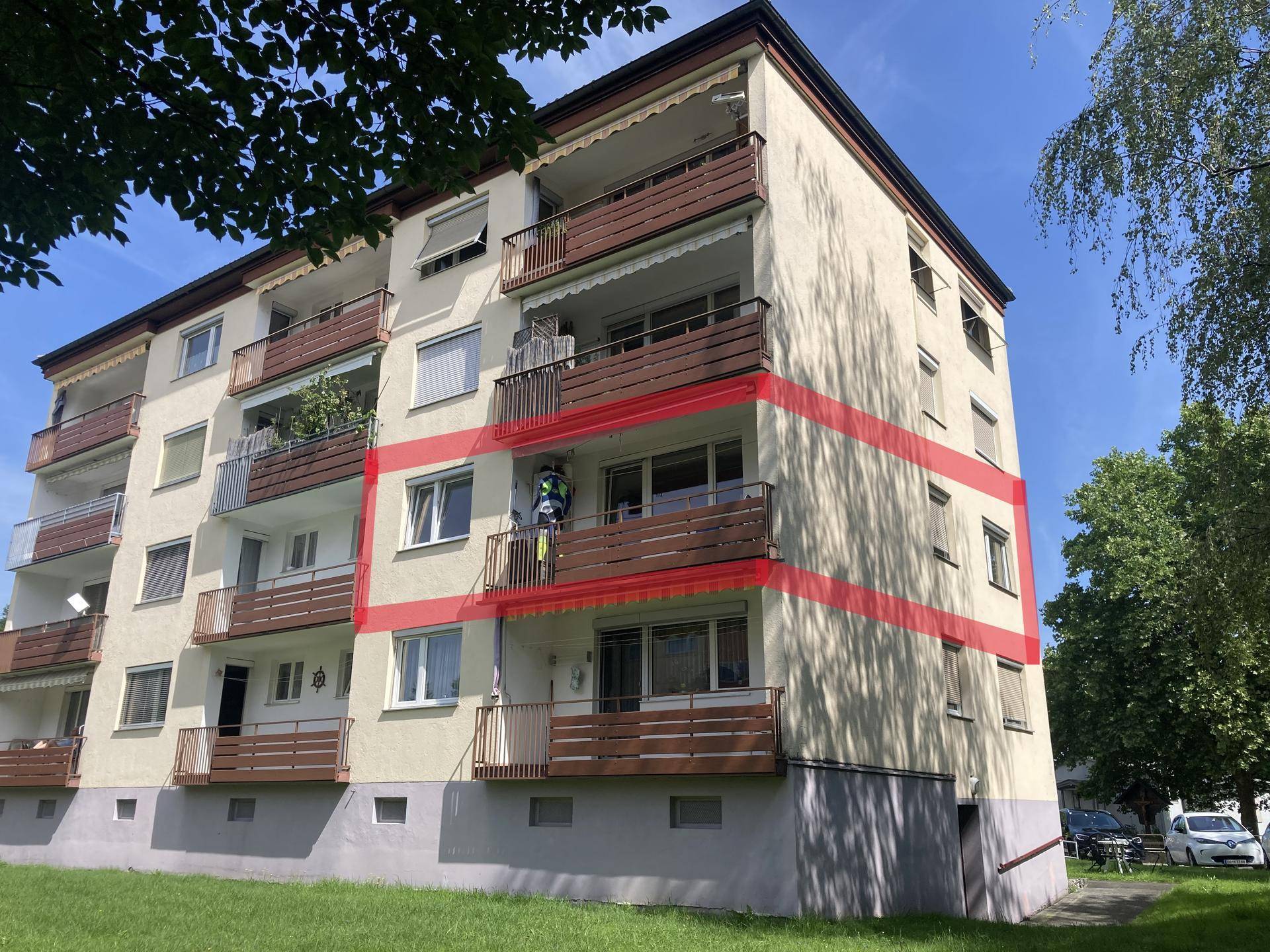 Mehrfamilienhaus mit mehreren Balkonen und grüner Umgebung unter blauem Himmel.