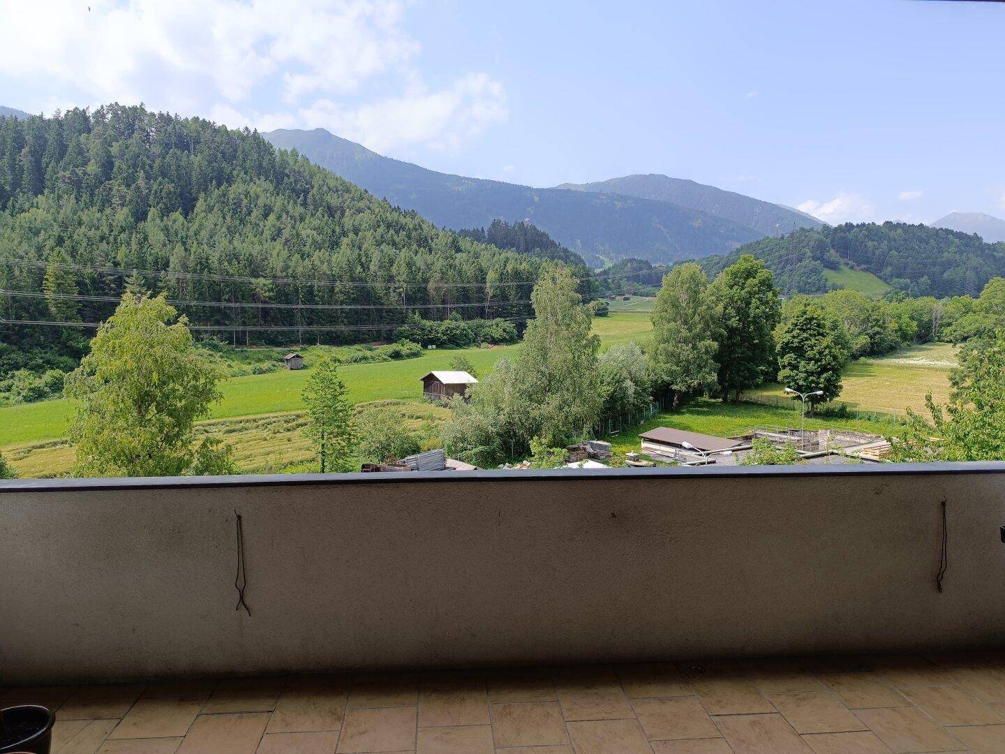 Balkon mit Fliesenboden und weitem Blick auf grüne Wiesen und bewaldete Berge unter blauem Himmel.