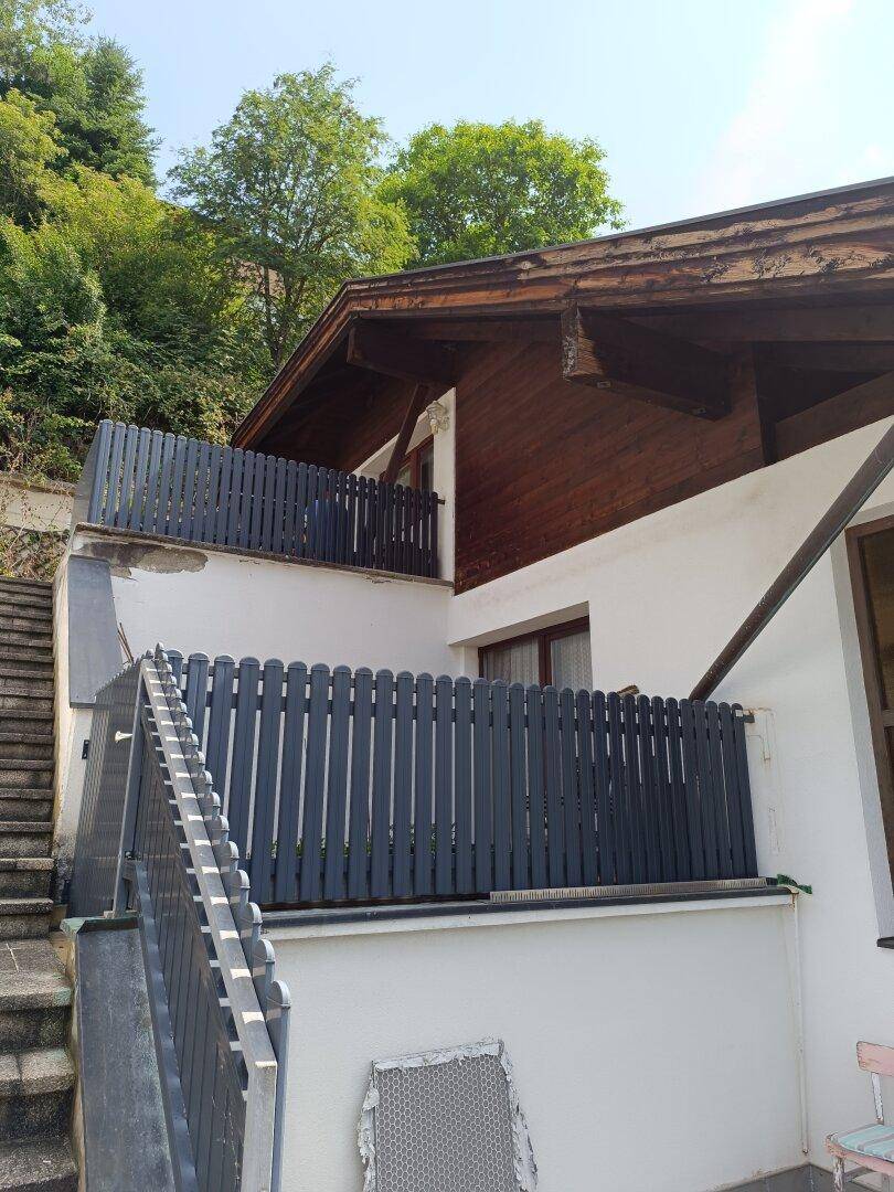 Seitenansicht des Hauses mit weißer Fassade, Holzelementen und grauen Balkongeländern sowie Außentreppen.