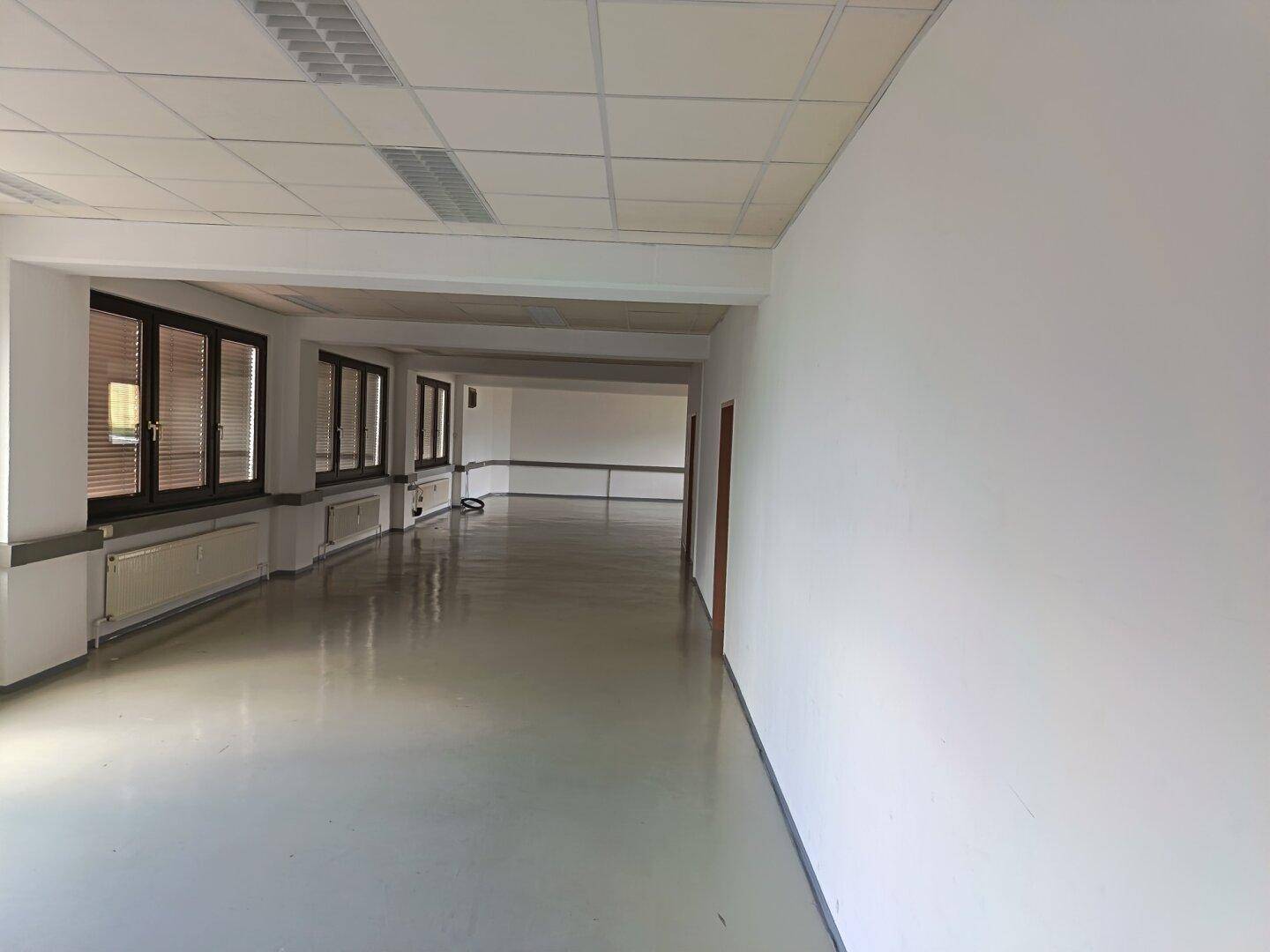 Langer, offener Bürobereich mit vielen Fenstern und glänzendem Bodenbelag, ideal für flexible Arbeitsplätze.