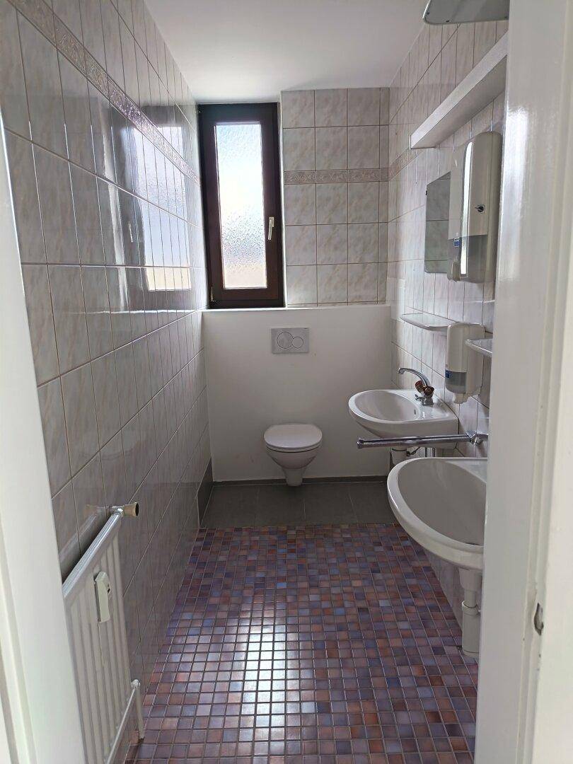 Kleines Badezimmer mit Fliesenboden, zwei Waschbecken, WC und einem Fenster für Tageslicht.