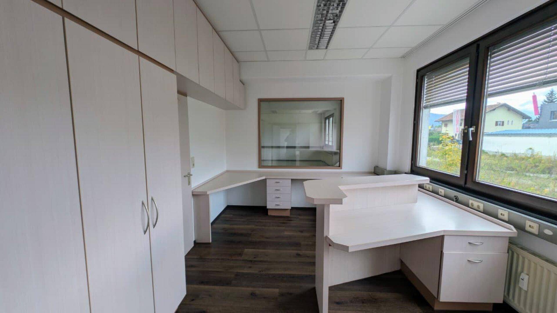 Büroraum mit L-förmigem Schreibtisch, Einbauschränken und Fenster mit Jalousien, bietet viel Arbeitsfläche.