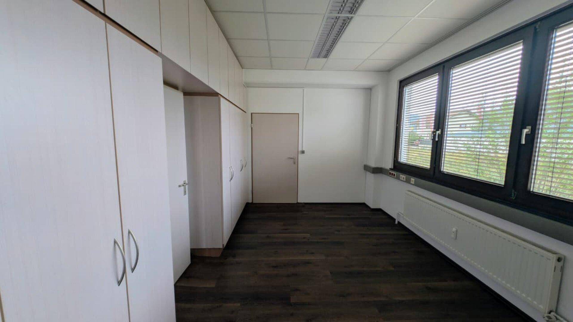 Helles Büro mit Einbauschränken, dunklem Boden und großen Fenstern mit Jalousien.