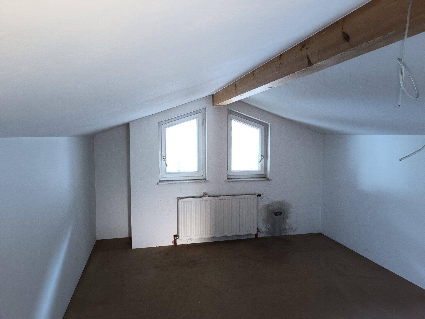 Helles Dachgeschosszimmer mit schrägen Wänden, sichtbaren Holzbalken und zwei Fenstern.