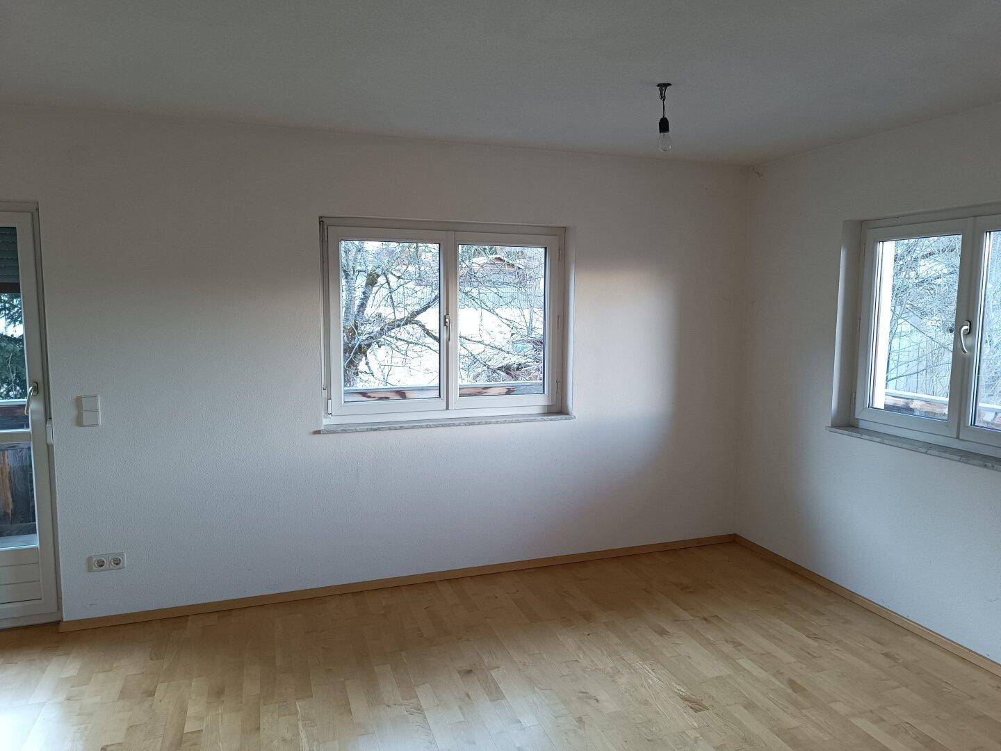 Heller Wohnraum mit Parkettboden und zwei Fenstern, die einen Blick nach draußen bieten.