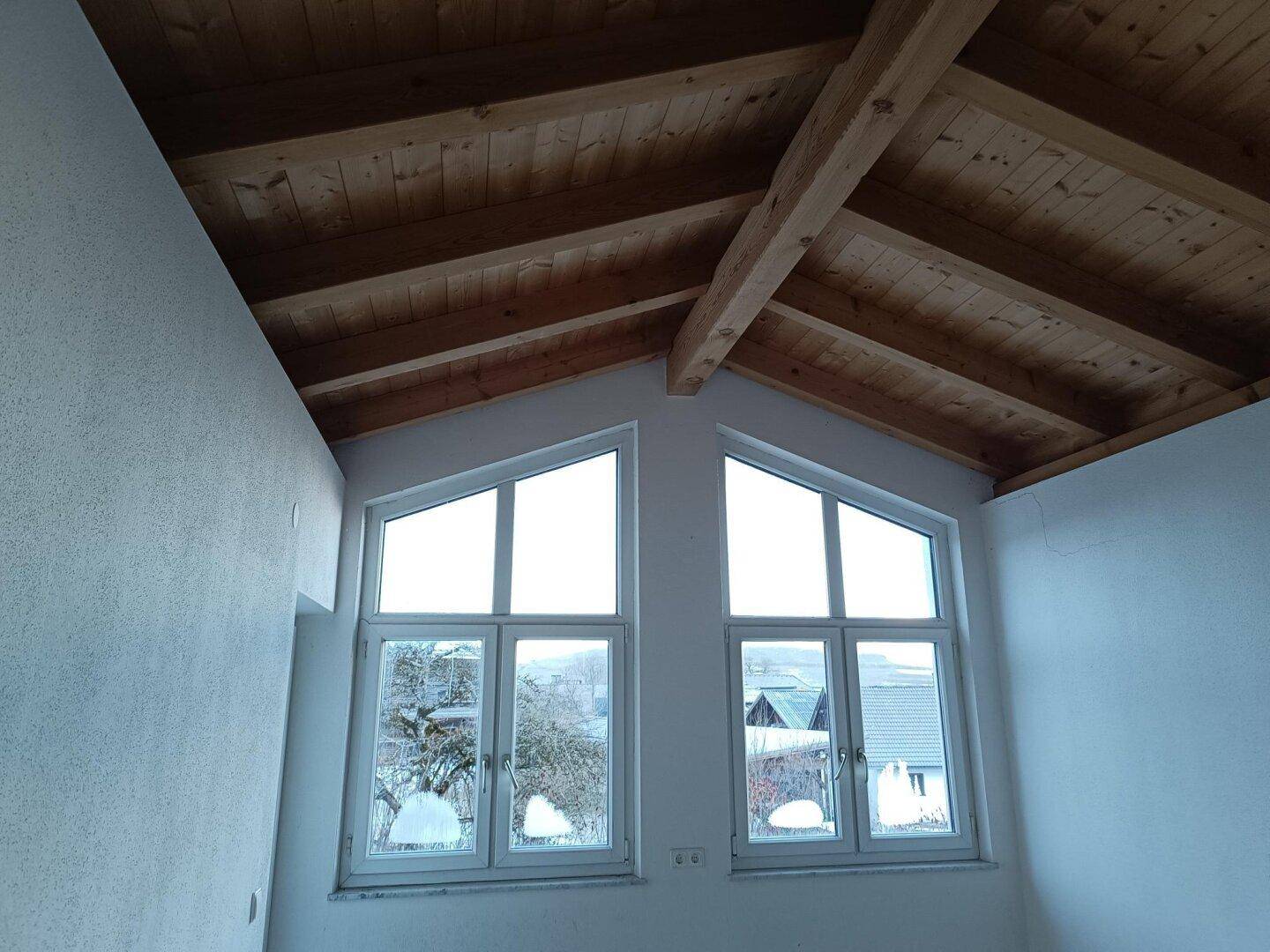 Dachgeschosszimmer mit sichtbarer Holzdecke, Holzbalken und zwei Giebelfenstern.