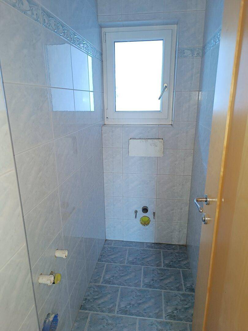 Kleines WC mit hellblauen Fliesen und einem Fenster, bereit für die Installation der Sanitärobjekte.