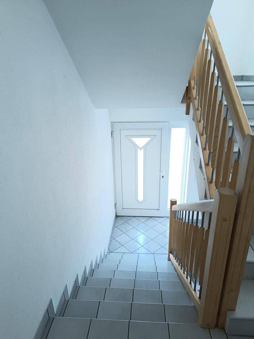 Treppenhaus mit Holzgeländer und gefliesten Stufen, führt zu einer Eingangstür.