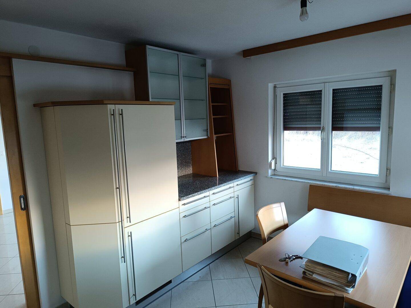 Helle Einbauküche mit ausreichend Stauraum, Essbereich und einem Fenster mit Rollladen.