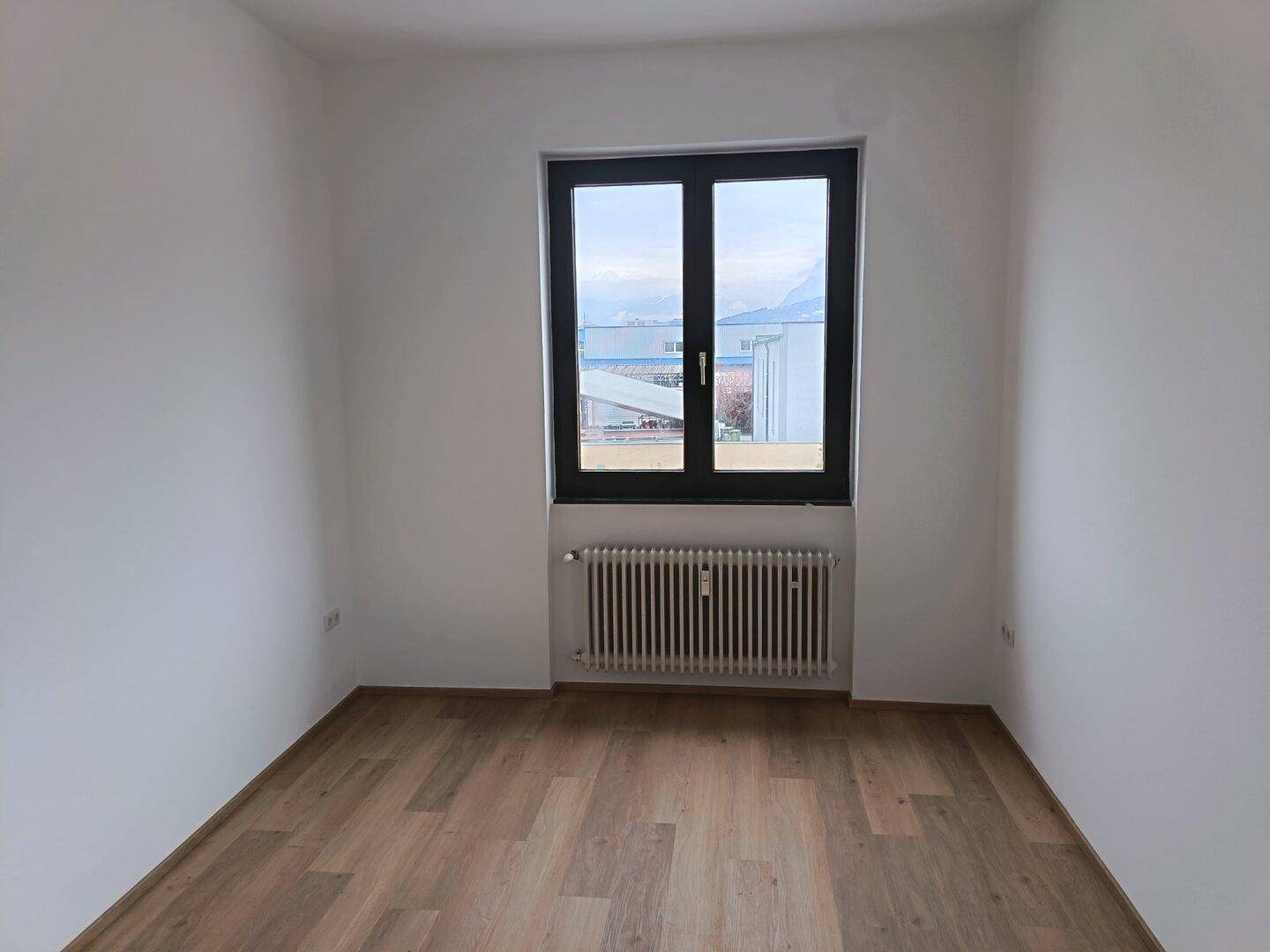 Helles Zimmer mit Holzboden, weißen Wänden und einem großen Fenster, ideal als Schlafzimmer.