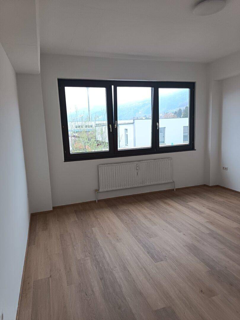 Geräumiges Zimmer mit Holzboden, weißen Wänden und großen Fenstern, ideal als Wohnzimmer.