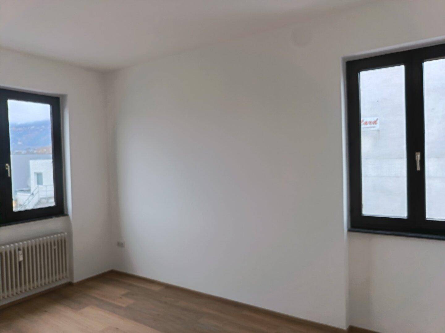 Helles Zimmer mit zwei Fenstern, Holzboden und weißen Wänden, bietet viel Tageslicht.