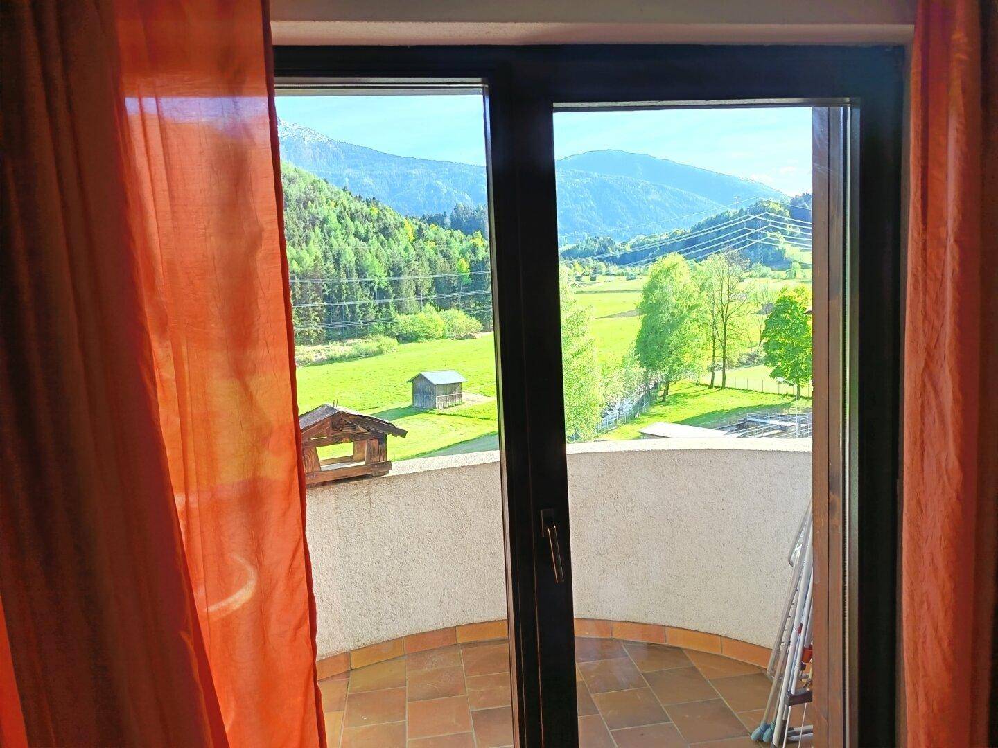 Blick aus dem Fenster auf einen Balkon und die weite grüne Landschaft mit Bergen im Hintergrund.