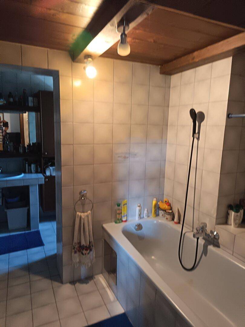 Badezimmer mit Badewanne, gefliesten Wänden und Holzdecke, bietet Zugang zu einem weiteren Raum.