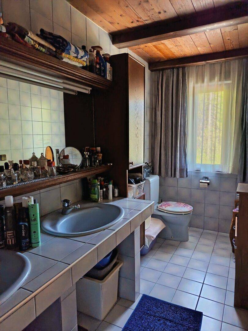 Badezimmer mit Doppelwaschbecken, WC, gefliesten Wänden und Holzdecke, gut beleuchtet durch ein Fenster.