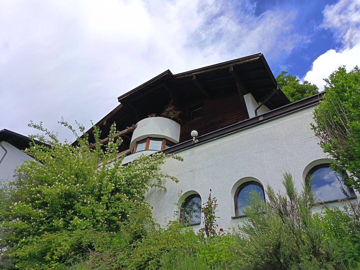 Weiße Hausfassade mit Rundbogenfenstern und Holzelementen, umgeben von üppigem Grün unter bewölktem Himmel.