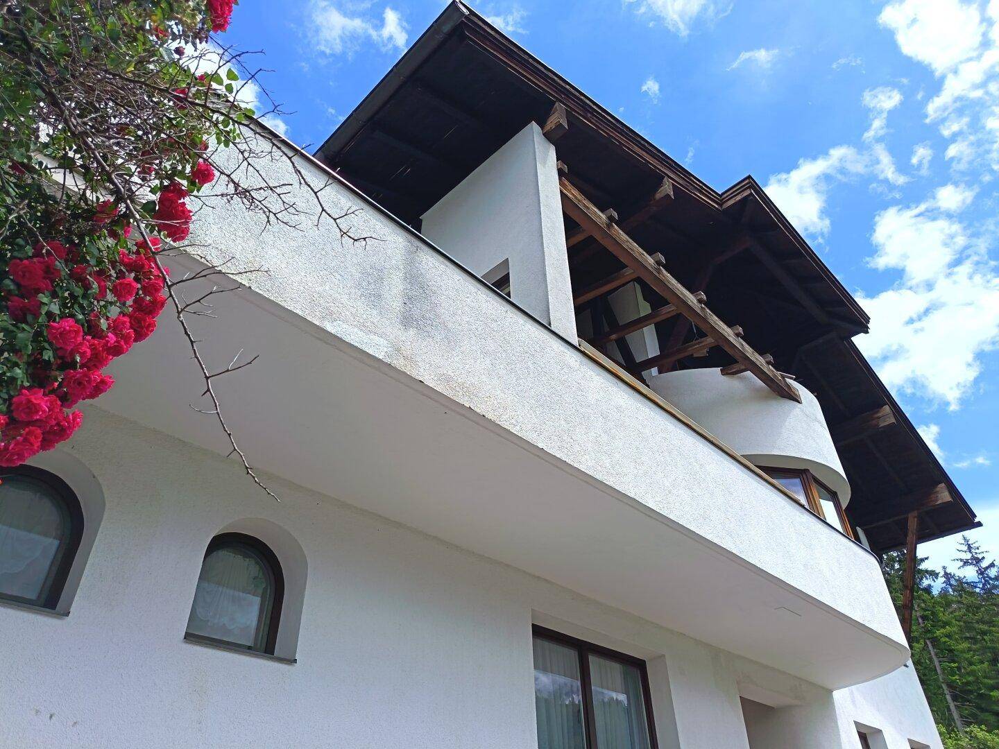 Detailansicht der weißen Hausfassade mit geschwungenem Balkon und roten Blumen unter blauem Himmel.