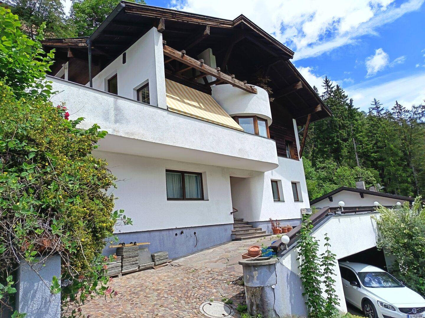 Mehrstöckiges Haus mit weißer Fassade, Holzbalkonen und einer Einfahrt mit Garage, umgeben von Bäumen.