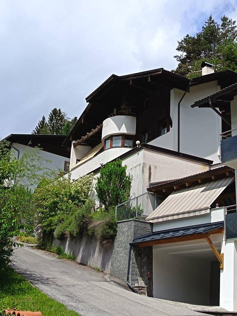 Hanglage des Hauses mit weißer Fassade, Holzbalkonen und einer Zufahrt, umgeben von viel Grün.