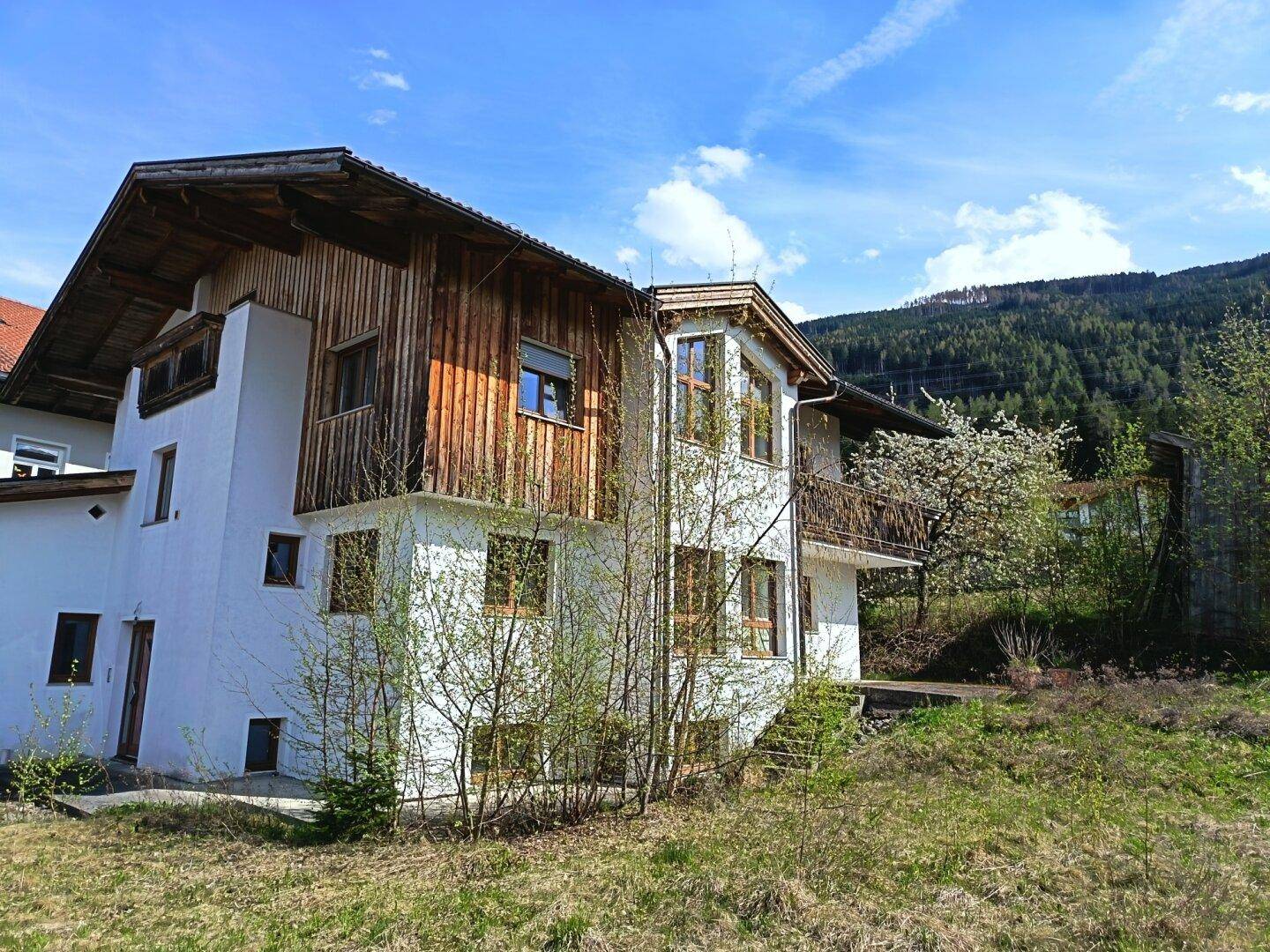 Wohnhaus mit weißer Fassade und Holzverkleidung, umgeben von Grünflächen und Bergen im Hintergrund.