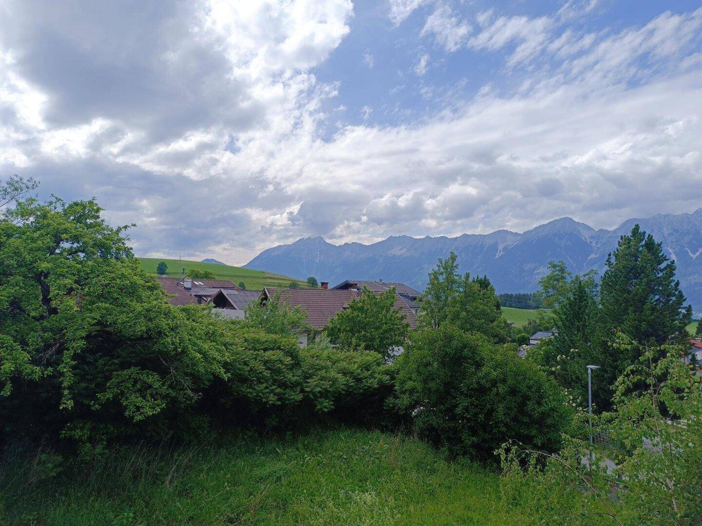 Panoramablick über grüne Baumwipfel und Dächer auf eine beeindruckende Bergkette unter bewölktem Himmel.