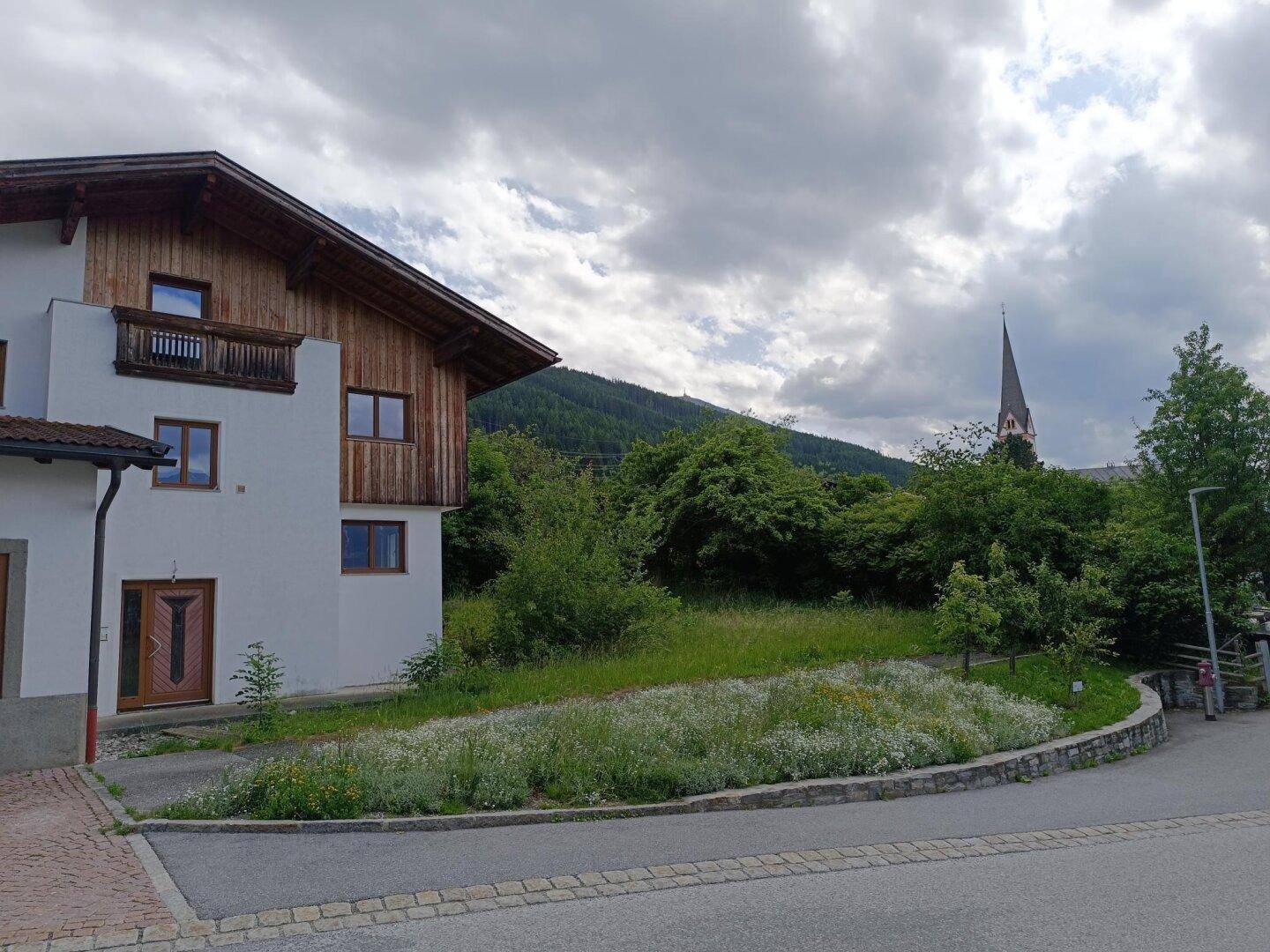 Seitenansicht des Wohnhauses von der Straße aus mit Blumenbeet und einem Kirchturm in der Ferne.