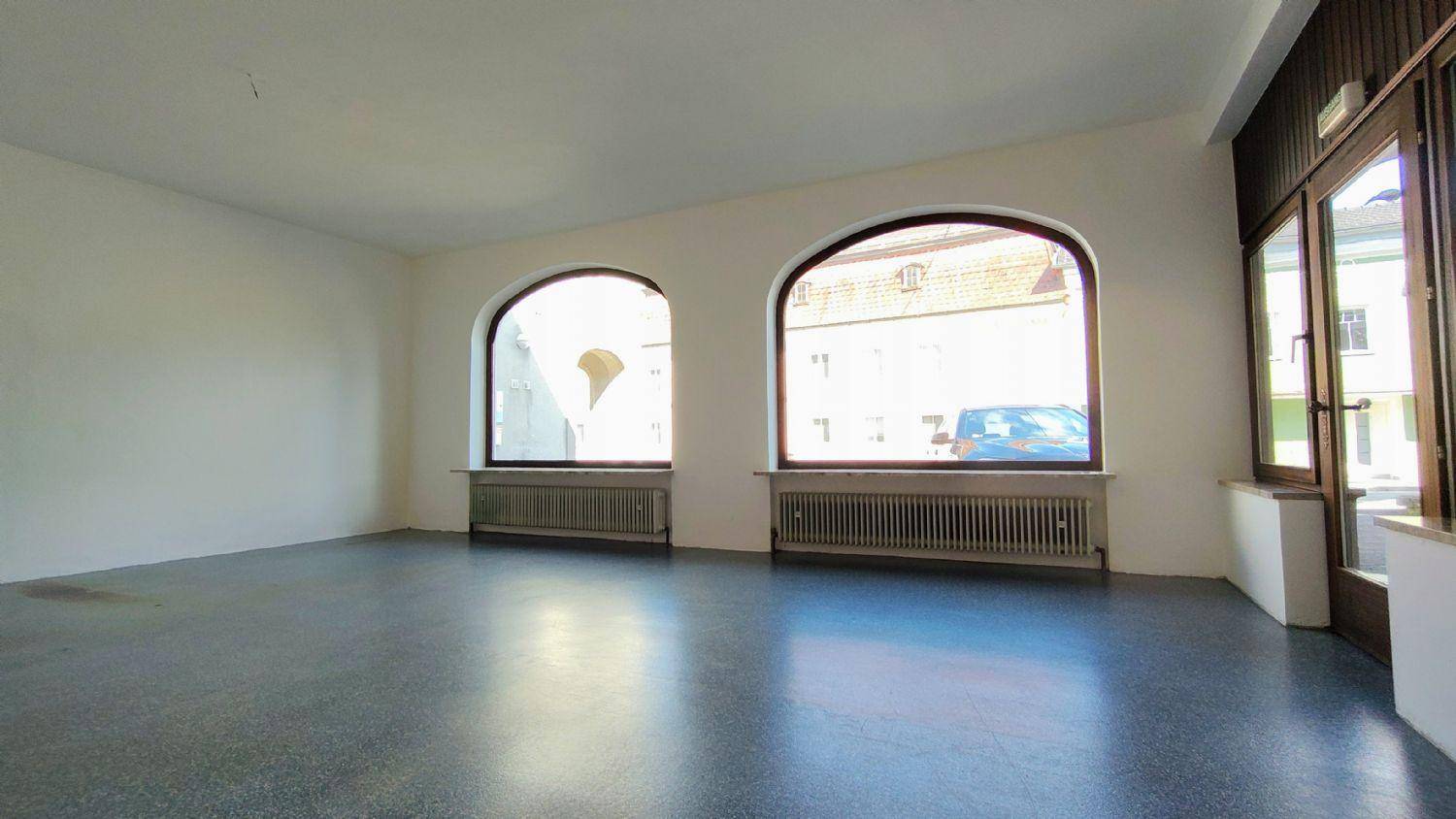 Geräumiger, leerer Raum mit hohen Bogenfenstern, die viel Tageslicht hereinlassen, und einem robusten Steinboden.