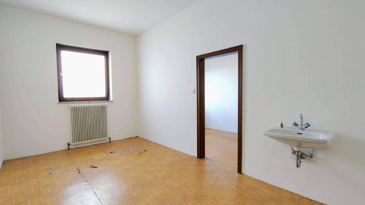 Einfacher Raum mit Waschbecken, Fenster und Heizkörper, der Bodenbelag weist deutliche Abnutzungsspuren auf.