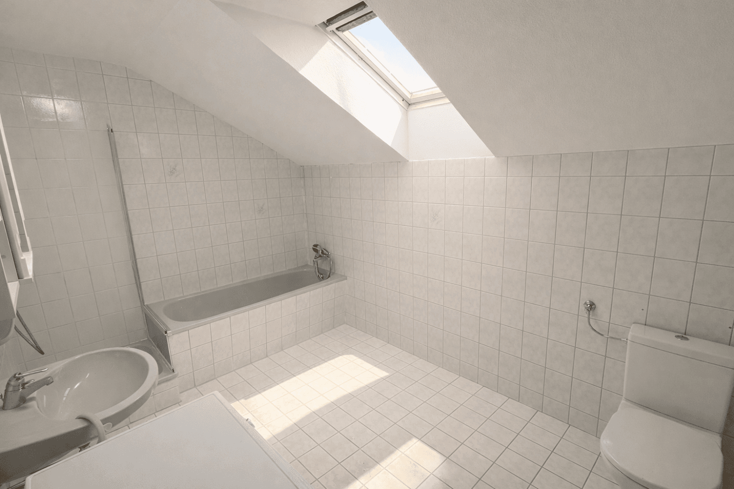 Helles Dachgeschoss-Badezimmer mit weißen Fliesen, Badewanne, Toilette und einem Dachfenster.