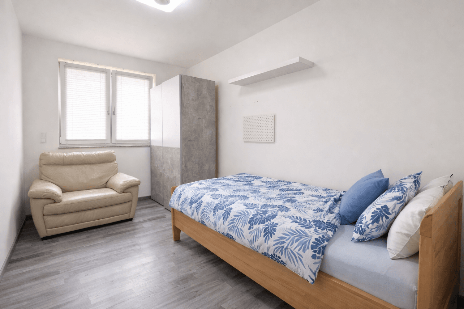 Helles Schlafzimmer mit Einzelbett, beigem Sessel und modernem Kleiderschrank auf Laminatboden.