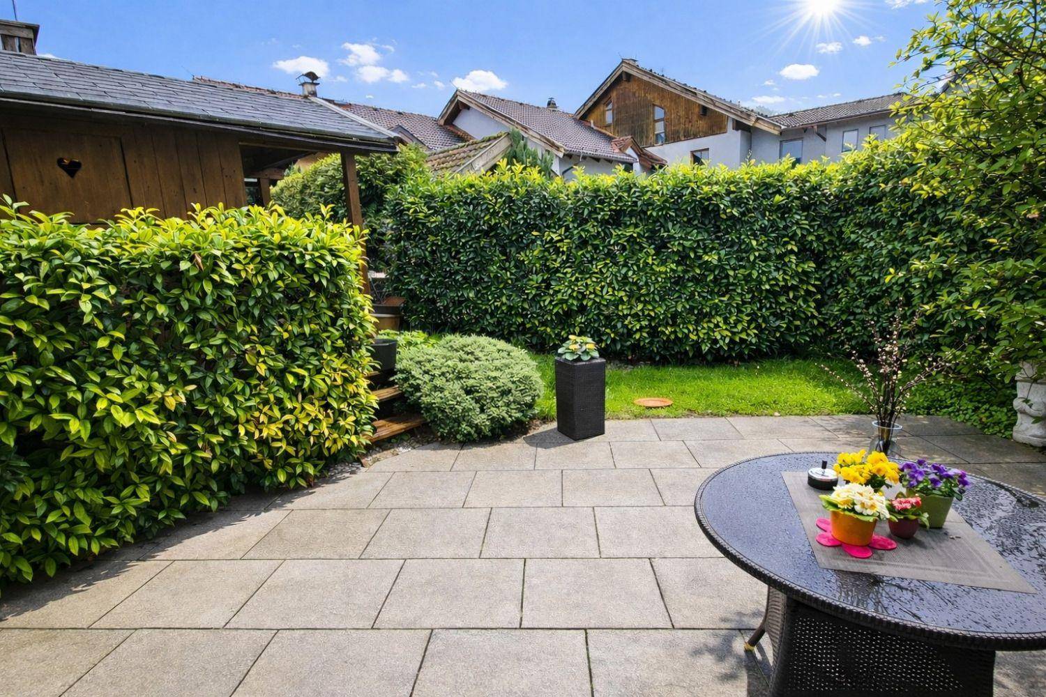 Helle Terrasse mit Gartentisch, Blumen und umgeben von einer grünen Hecke.
