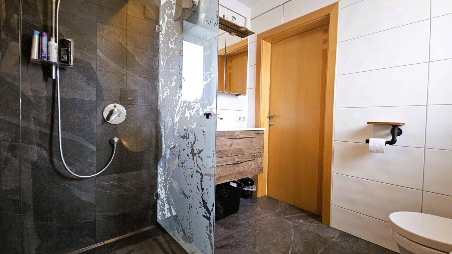 Modernes Badezimmer mit begehbarer Dusche, dunklen Fliesen und Holzwaschtisch.
