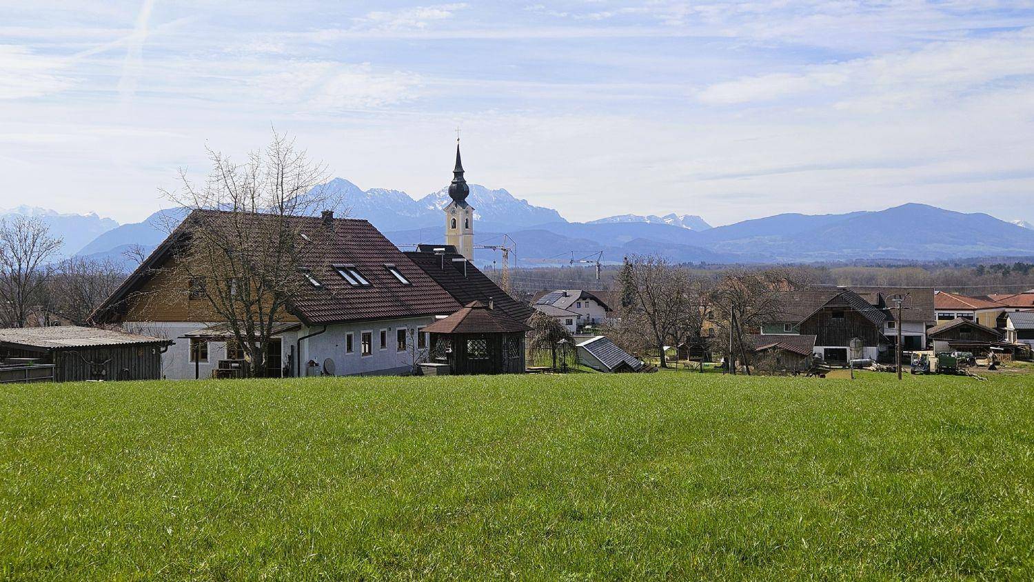 Ländliche Szenerie mit traditionellen Häusern, einer Kirche und einer beeindruckenden Bergkette im Hintergrund.