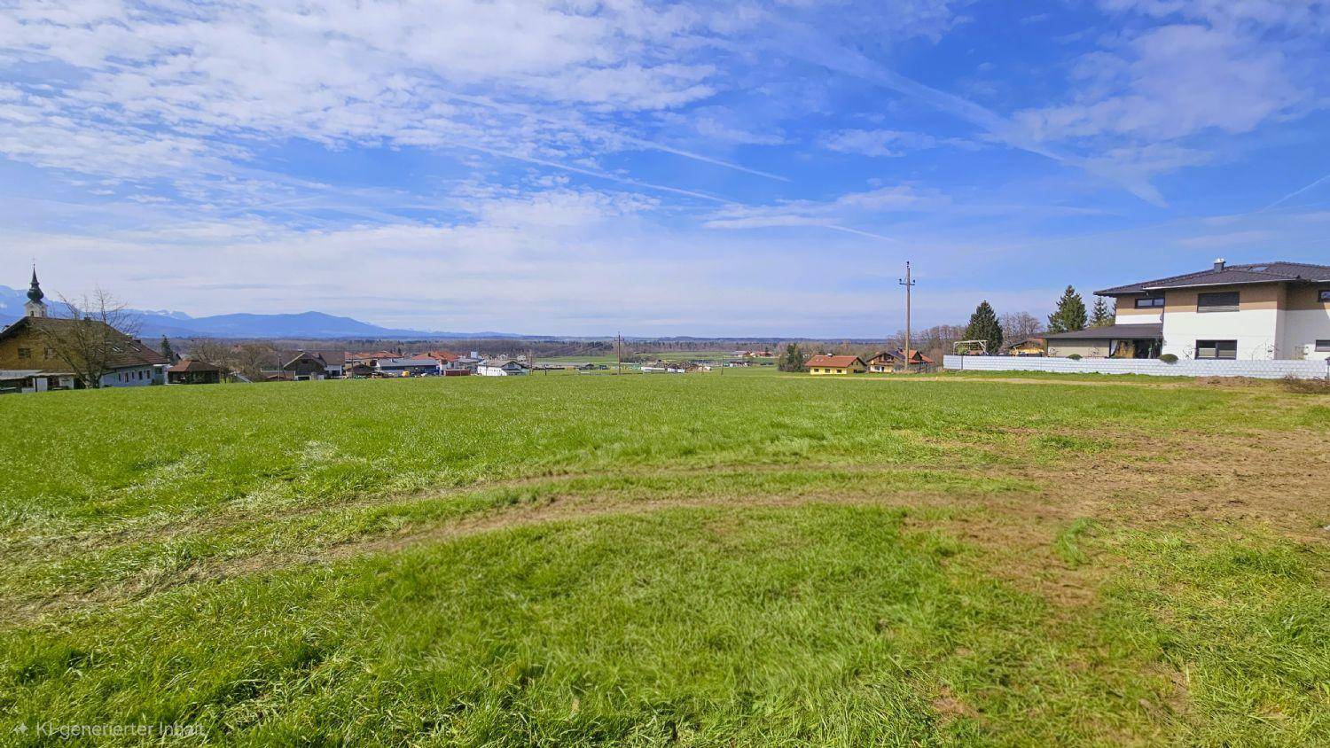 Weitläufiges grünes Feld mit Blick auf eine ländliche Siedlung und Berge am Horizont unter blauem Himmel.