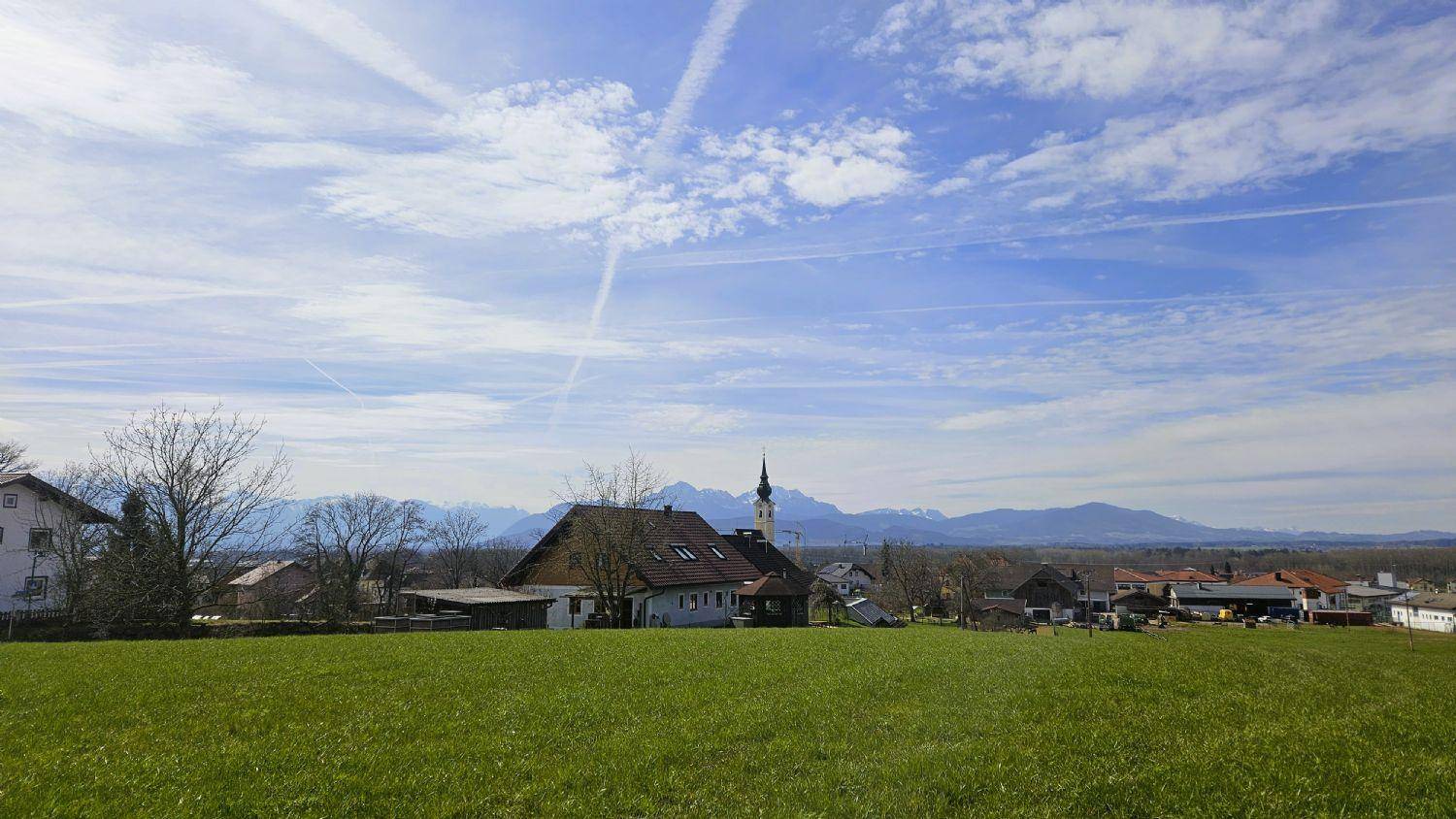 Weitläufige grüne Landschaft mit ländlicher Bebauung und majestätischer Bergkette im Hintergrund unter blauem Himmel.