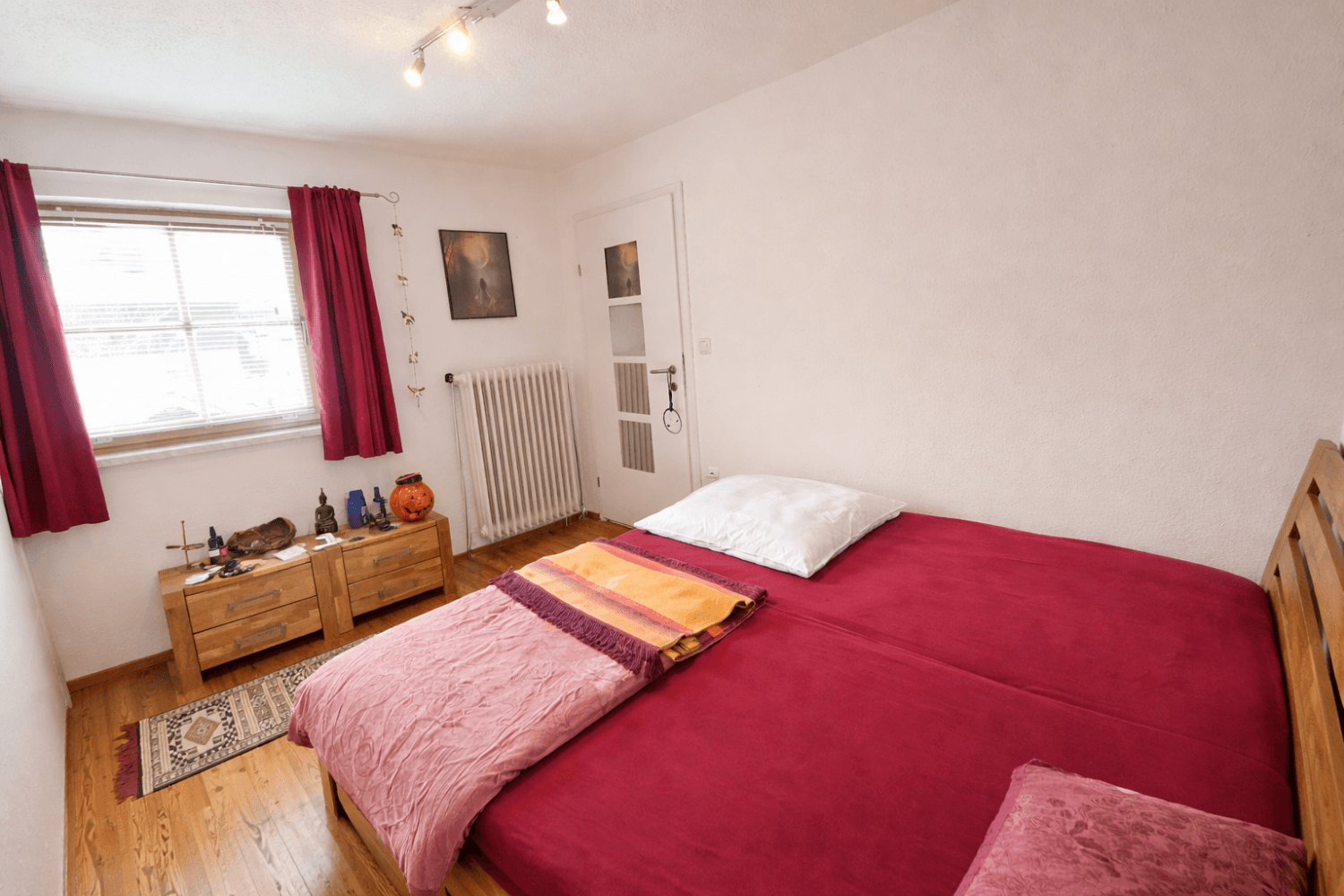 Schlafzimmer mit Holzbett, Holzboden und roten Vorhängen, gut beleuchtet.