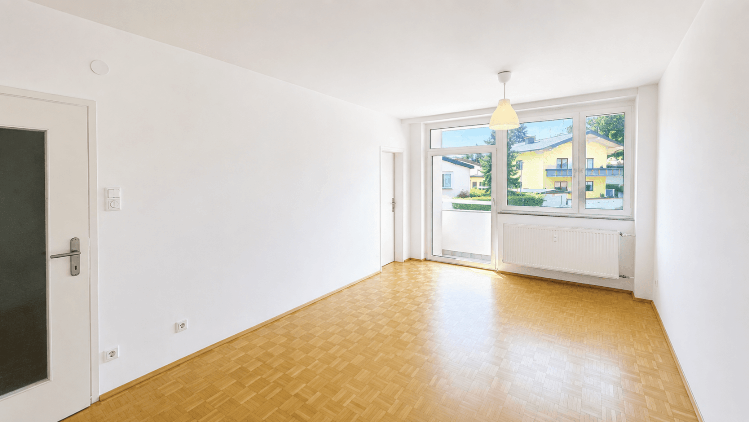 Helles, leeres Wohnzimmer mit Parkettboden, weißen Wänden und Zugang zu einem Balkon.