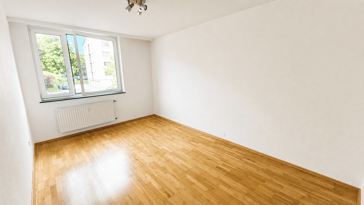 Leeres Schlafzimmer mit hellem Parkettboden, weißen Wänden und einem großen Fenster.