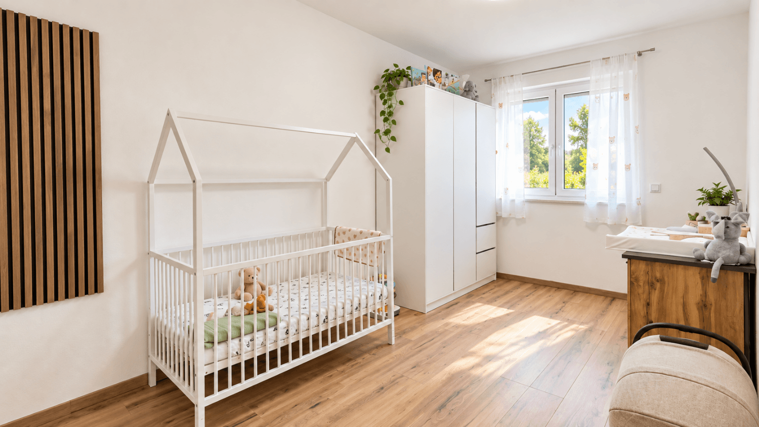 Helles Kinderzimmer mit weißem Gitterbett, Kleiderschrank und Wickeltisch am Fenster.