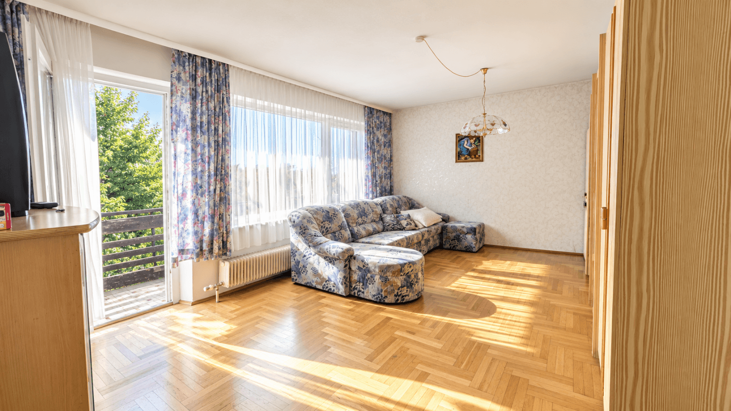 Helles Wohnzimmer mit Fischgrätparkett, gemustertem Sofa und großem Fenster mit Zugang zum Balkon.