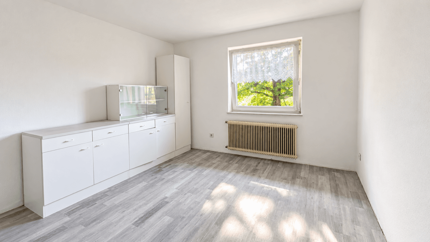 Leerer Raum mit hellgrauem Laminatboden, weißen Wänden und einem Fenster mit Spitzenvorhang.
