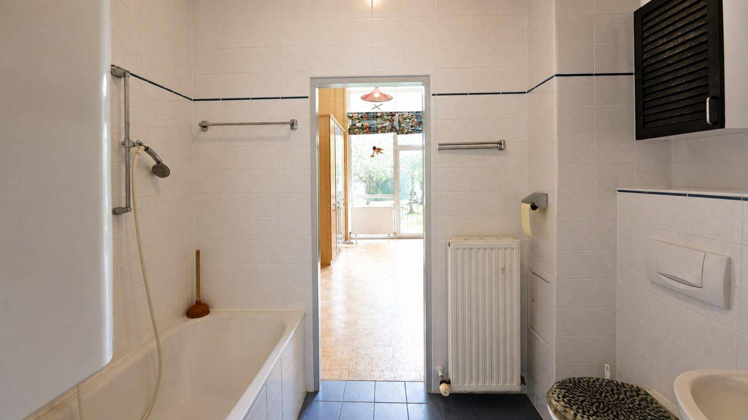 Älteres Badezimmer mit weißen Fliesen, Badewanne und Blick in den angrenzenden Raum.